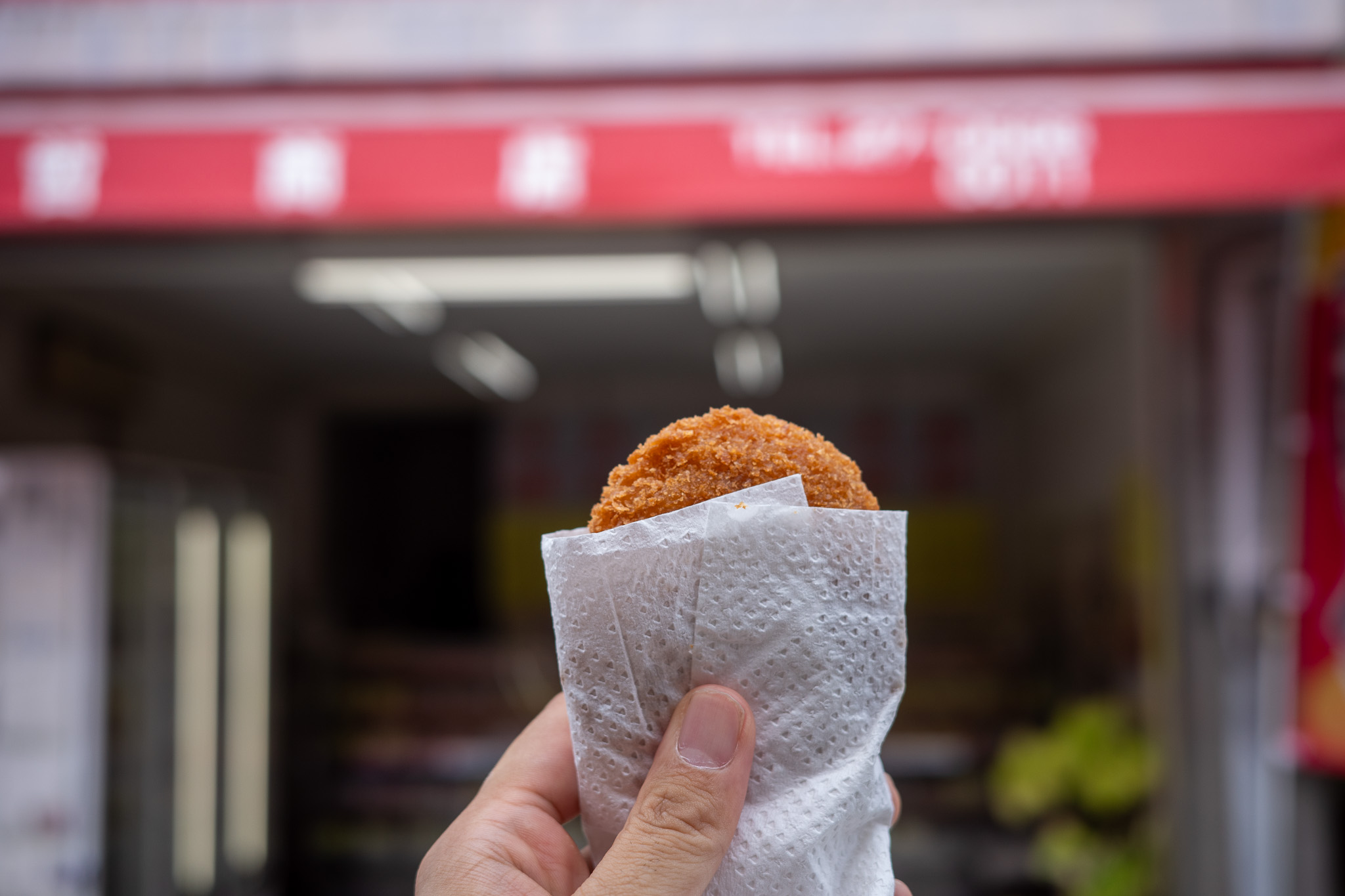 手に持ったコロッケ、背景に赤い店舗看板